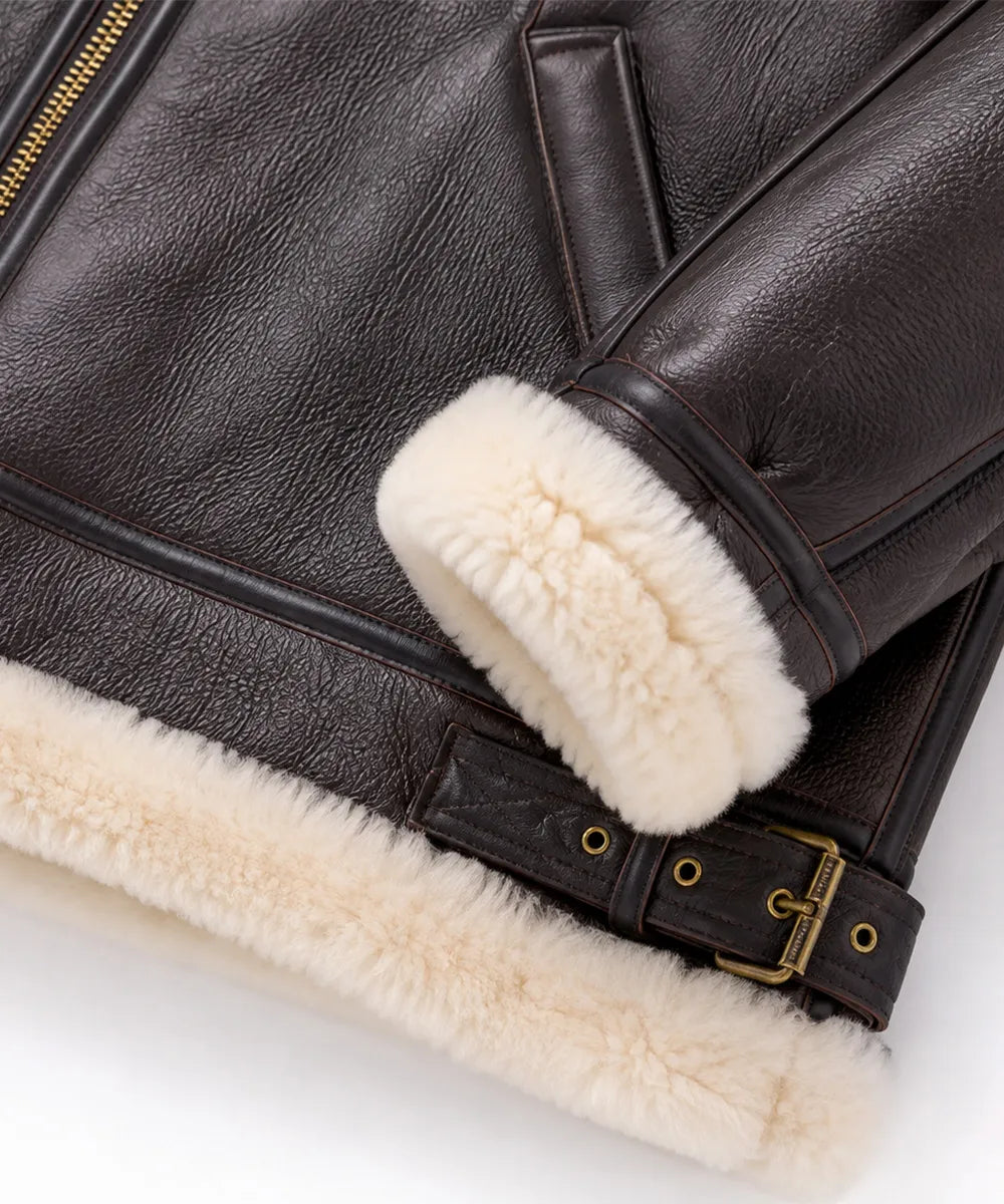 Close-up of a dark brown leather jacket with thick cream shearling lining on the cuffs and hem, featuring a gold-tone buckle detail.