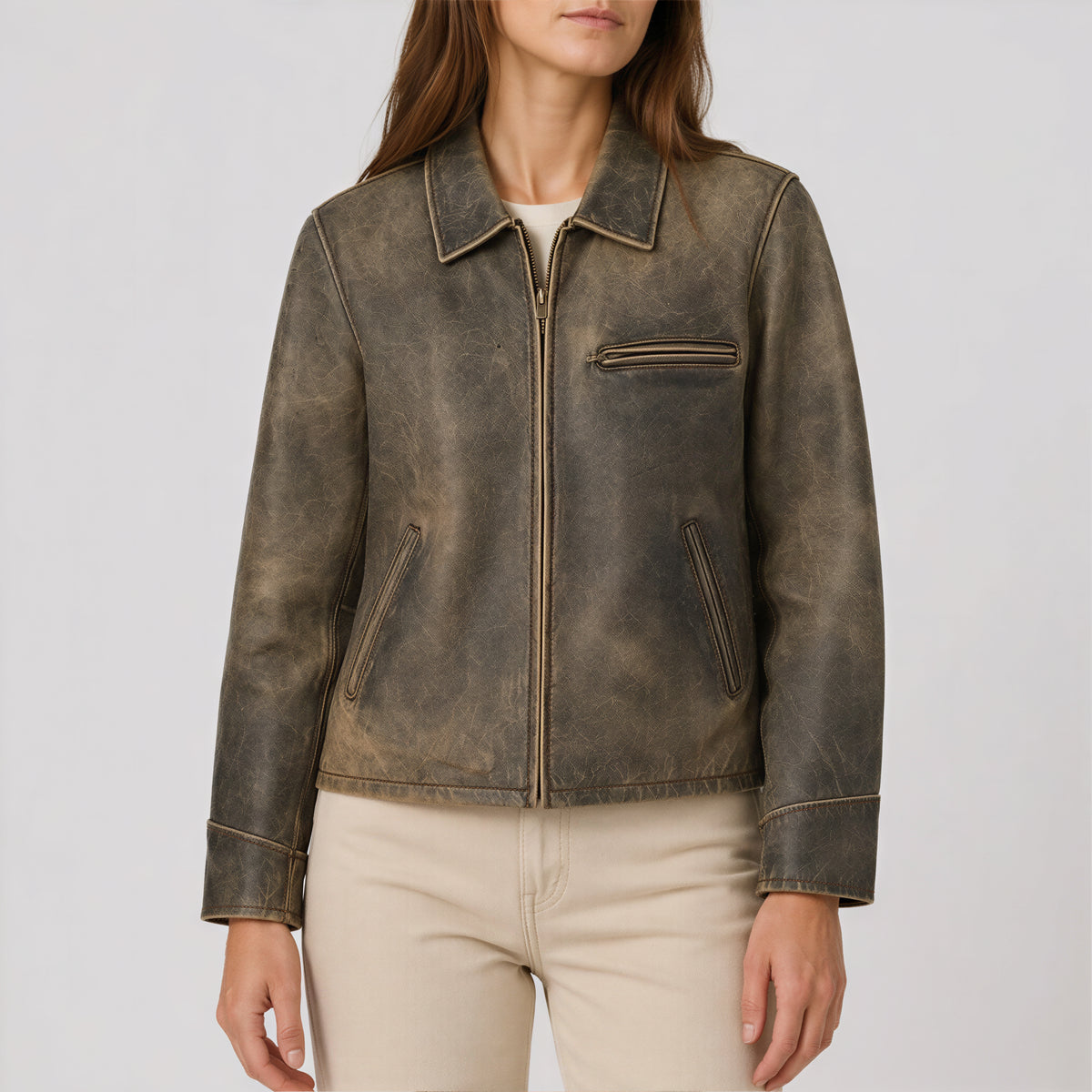 A woman wearing a distressed brown leather jacket with a front zipper, collar, and zippered chest pocket, paired with light beige pants, against a plain light gray background.