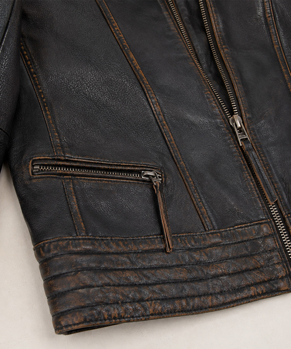 Close-up of a distressed black leather jacket showing the texture, contrast stitching, a zippered pocket, and ribbed cuffs on a light neutral background.