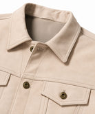 Close-up of a light beige canvas jacket with a pointed collar, buttoned front, and flap chest pockets with brass-tone buttons.