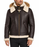 A man wearing a dark brown leather aviator-style jacket with thick cream shearling lining on the hood, collar, cuffs, and hem, paired with black pants against a white studio background.