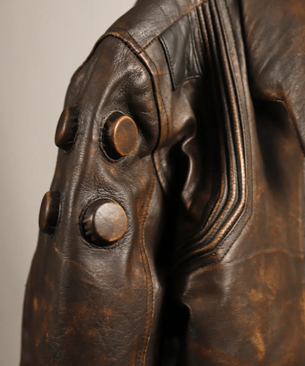 Close-up of a distressed brown leather jacket featuring large round metallic buttons on the shoulder and sleeve, with detailed stitching and a textured leather surface.