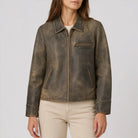 A woman wearing a distressed brown leather jacket with a front zipper, collar, and zippered chest pocket, paired with light beige pants, against a plain light gray background.