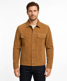 A man wearing a tan suede jacket over a white t-shirt and dark blue jeans, standing against a plain white studio background.