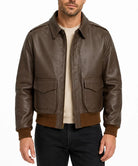 A man wearing a brown leather bomber jacket with a tan crewneck sweater and black jeans, photographed against a plain white background.
