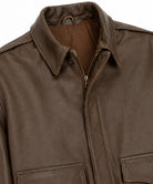 Close-up of a brown leather jacket showing the collar, zipper, and front pockets against a white background.