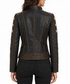 A woman viewed from the back wearing a black leather jacket with decorative gold-toned stitching, lace-up details, and star motifs on the sleeves, paired with black jeans against a plain white background.