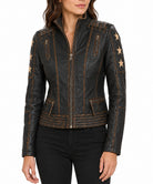 A woman wearing a black distressed leather moto jacket with gold-toned star details on the sleeves and gold contrast stitching, paired with black pants against a white background.