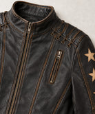Close-up of a dark brown distressed leather jacket with gold-toned zippers, contrast stitching, and gold star patches on the sleeve, shown against a light textured background.