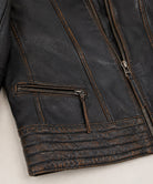 Close-up of a distressed black leather jacket showing the texture, contrast stitching, a zippered pocket, and ribbed cuffs on a light neutral background.