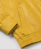 Close-up of a mustard yellow leather jacket showing the sleeve cuff, elasticated hem, and a vertical welt pocket on a light gray background.